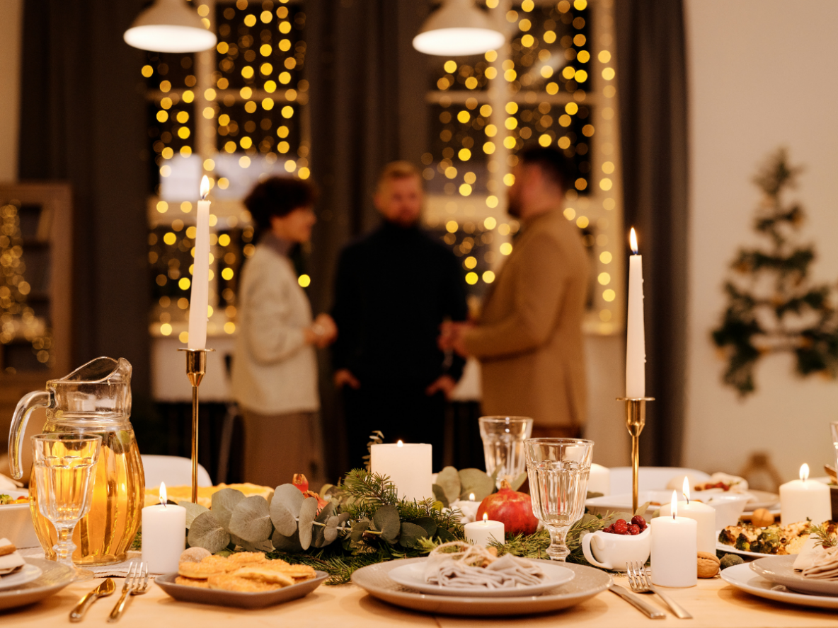 A table with plates, glasses, food and candles with three people standing in the background talking